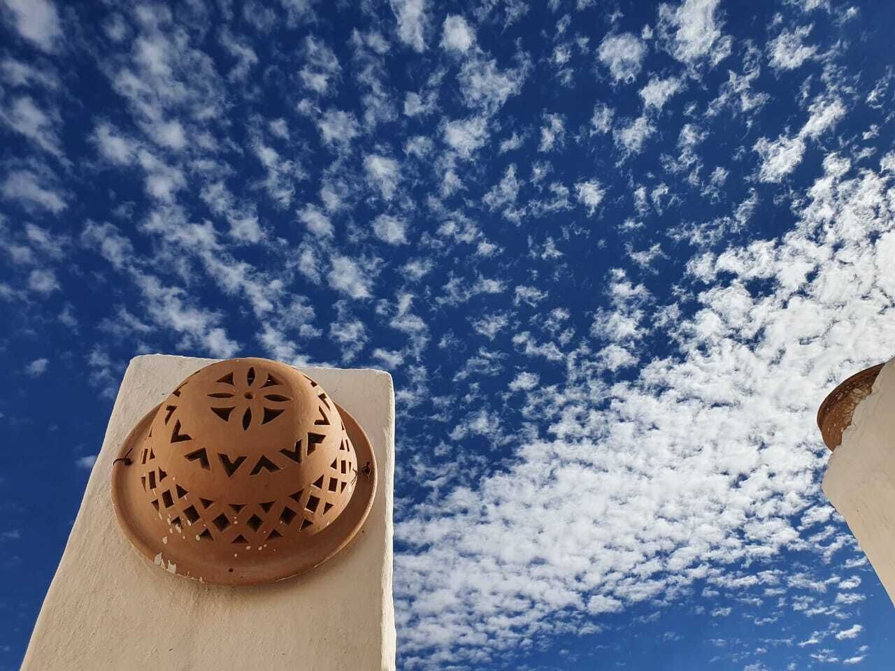 En lerpotte med geometriske mønstre på en hvid væg, mod en blå himmel med spredte skyer.