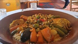 A large, round, brown serving dish filled with a variety of vegetables and legumes, placed on a table in a room with white walls and framed artwork. - DAR INFIANE Tata  (Tigzmerte)
