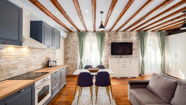 A modern kitchen with a stone backsplash, wooden beams, a dining area with a table and chairs, and a living area with a sofa. - Stephane City Vibe Suites (Trogir)