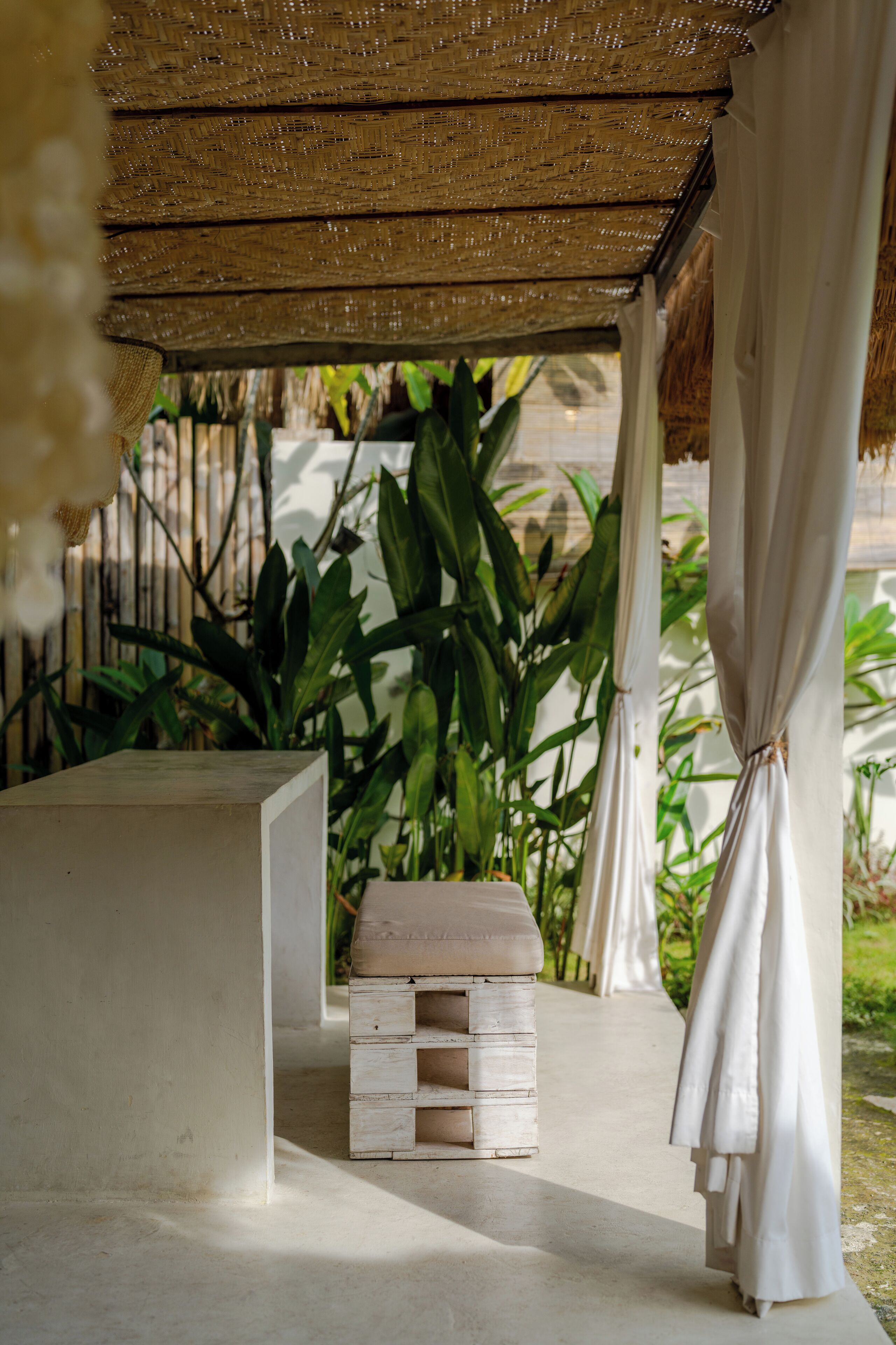 A covered outdoor seating area with a thatched roof, a white bench, and lush green plants in the background.