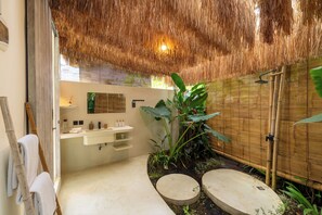 Une salle de bain avec un plafond en chaume, un lavabo blanc, un miroir et un espace douche.