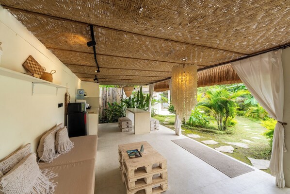 Une pièce dotée d’un plafond en fibres tressées, d’un canapé agrémenté de coussins en macramé, d’une table basse en bois et offrant une vue sur un jardin avec un élément décoratif aquatique.