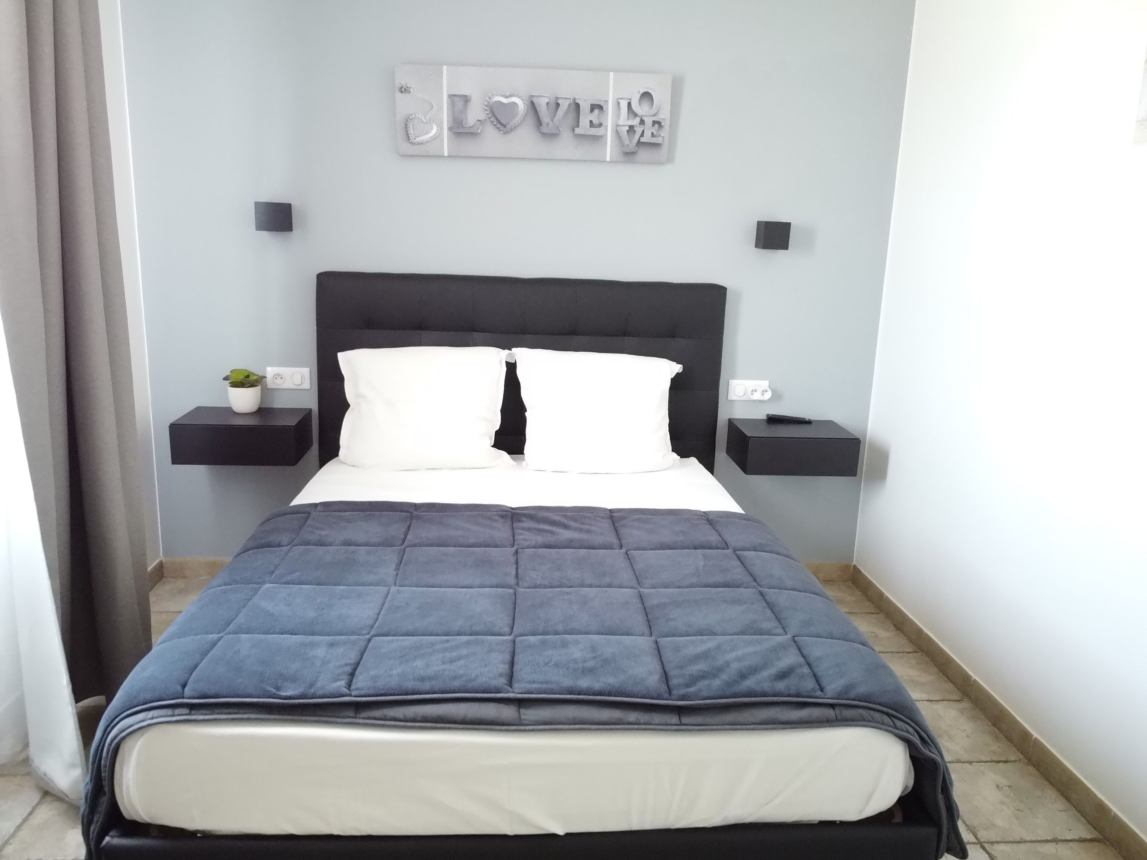 Ein ordentlich bezogenes Bett mit einer dunkelblauen Tagesdecke, weißen Kissen und einem schwarzen Kopfteil. Auf einem Nachttisch steht eine kleine Pflanze und eine an der Wand befestigte Schilder mit der Aufschrift "LOVE".