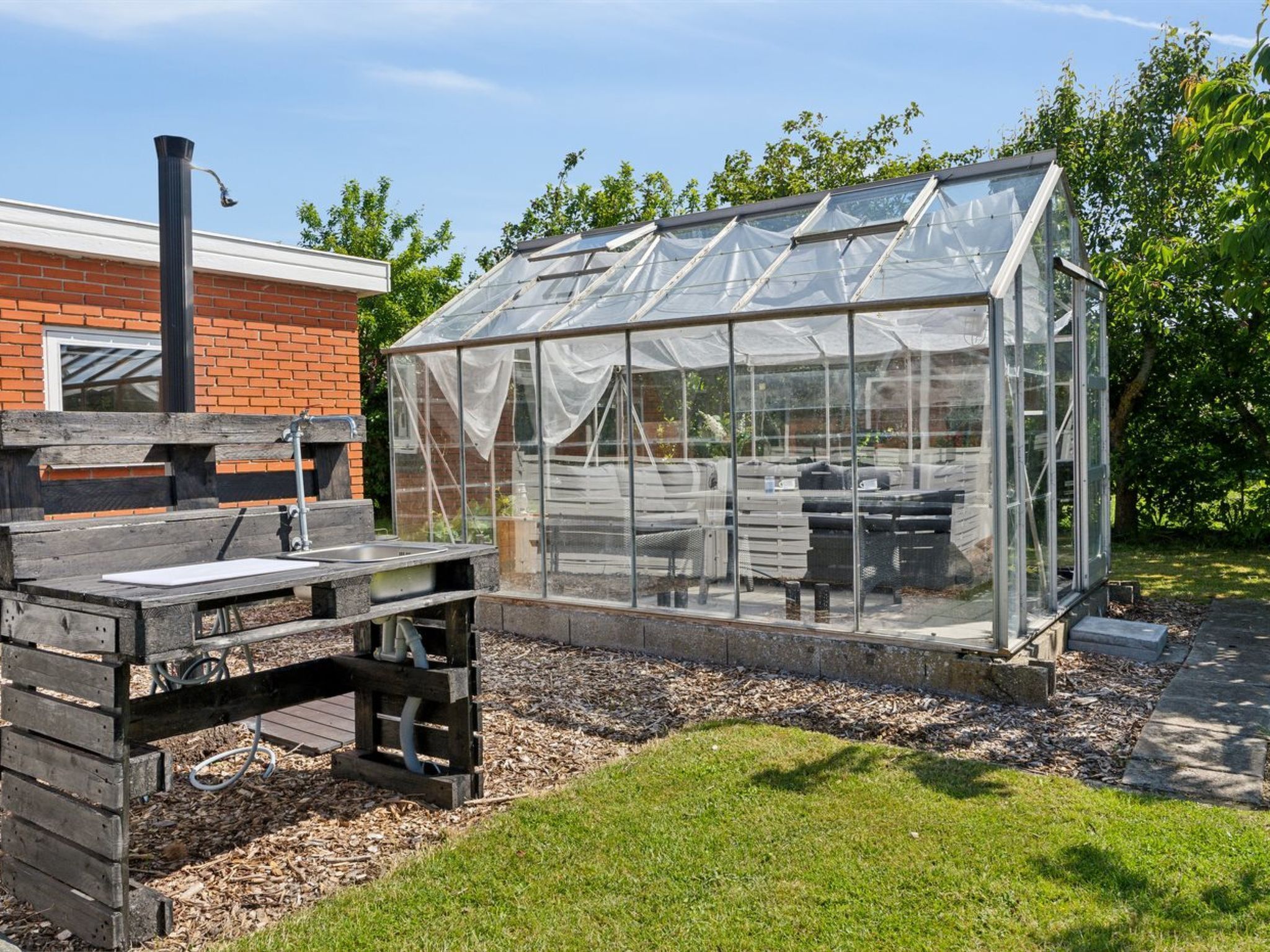 A greenhouse with a transparent roof, a wooden table, and a brick building in the background.