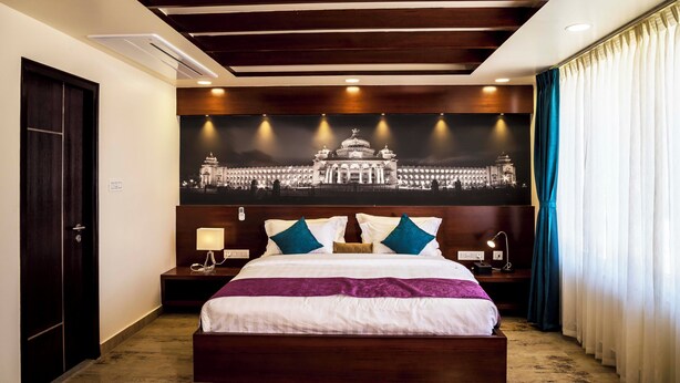 A hotel room with a large bed, a nightstand with a lamp, a wall-mounted TV, and a large framed picture of a building. - Hotel Urban Jade (Bengaluru)