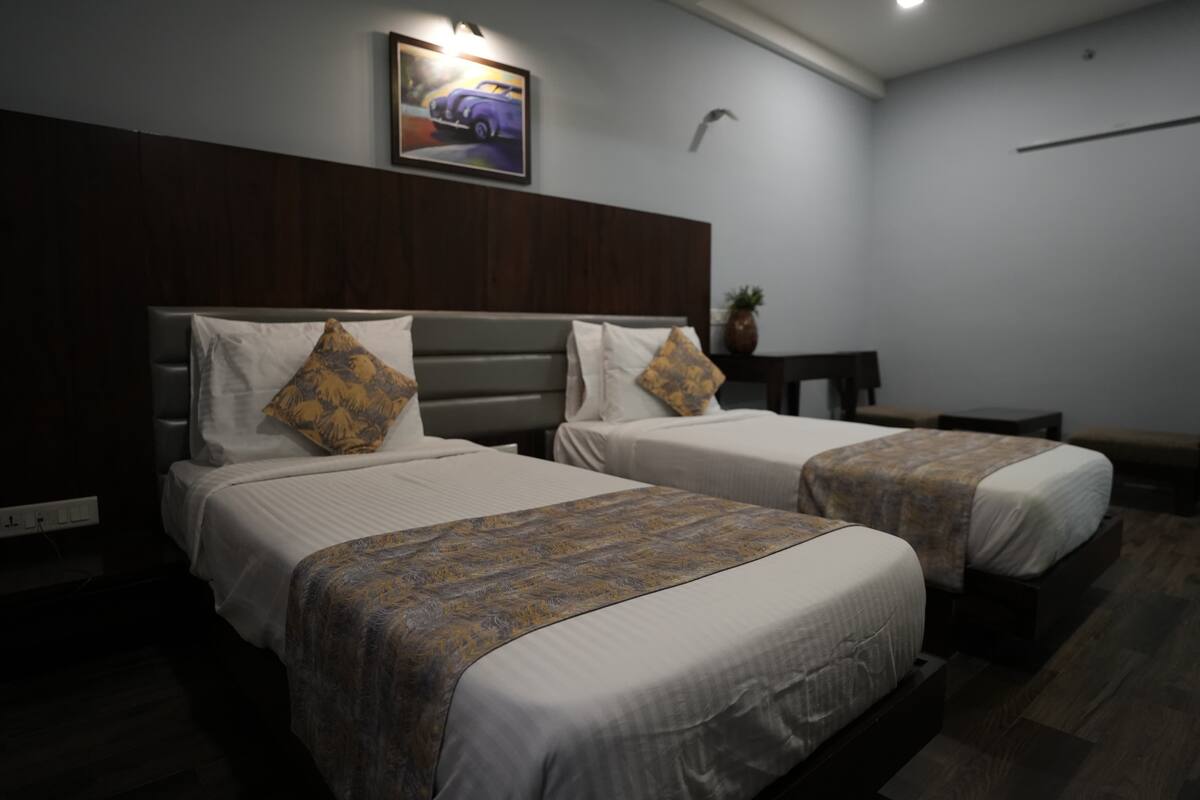 A hotel room with two beds, each with a headboard and pillows. There is a painting on the wall above the beds and a small table with a plant beside the window.