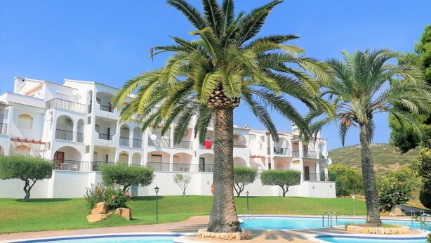 Un resort con área de piscina, palmeras y un edificio blanco con balcones.