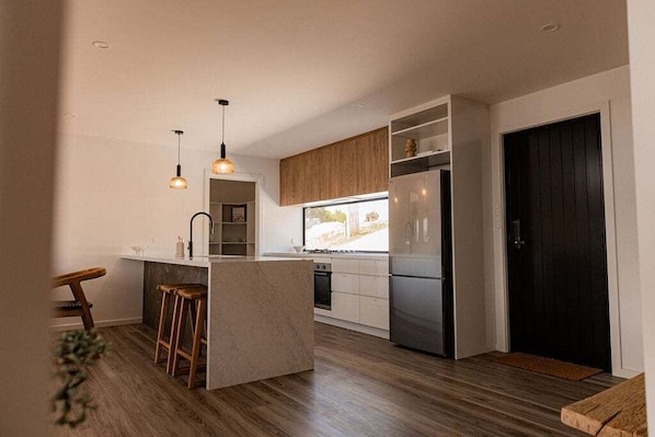 A modern kitchen with wooden cabinets, a central island, bar stools, pendant lighting, and a black door.