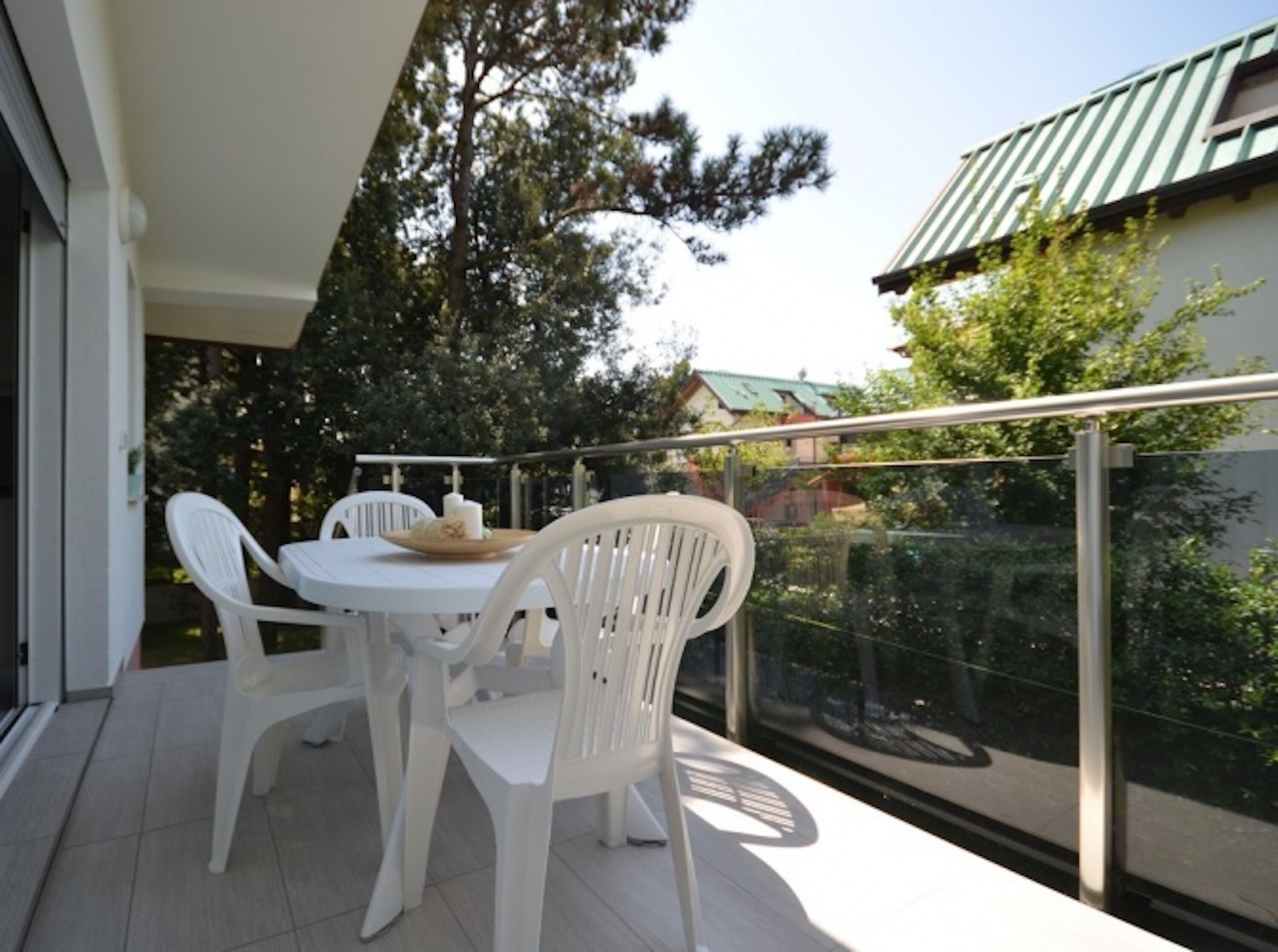 A balcony with a white table and chairs, a glass railing, and a view of trees and a building.