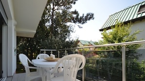 A balcony with a white table and chairs, a glass railing, and a view of trees and a building.