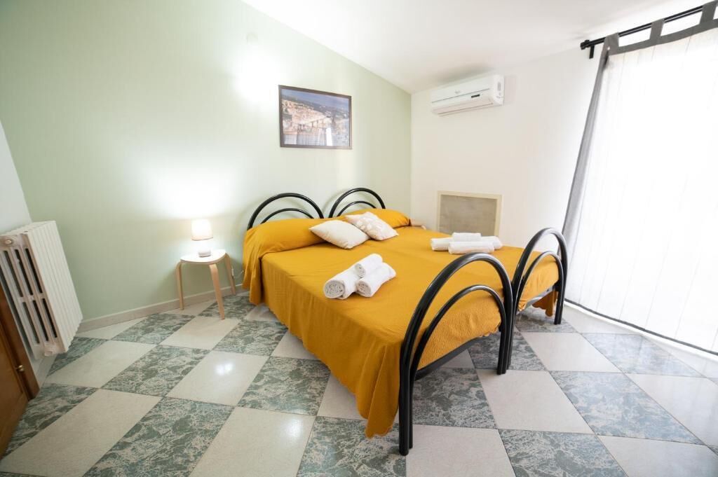 A room with a bed, a small table with a lamp, a radiator, and a window with curtains.
