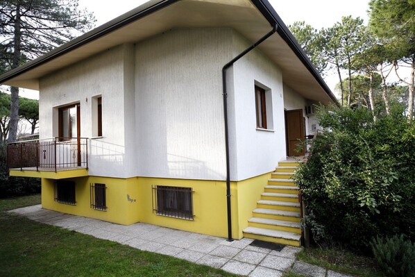 Une maison à deux étages, à la façade jaune et blanche, dotée d’un balcon avec une rambarde en métal et d’un escalier menant à l’entrée.