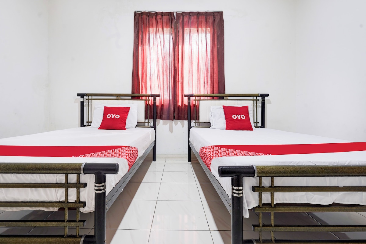 A hotel room with two single beds, each with a red and white bedspread and matching pillows. The room has white walls and a tiled floor.
