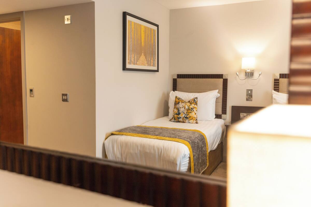 Una habitación de hotel con una cama, un cuadro en la pared, una mesita de noche y una lámpara fijada en la pared.