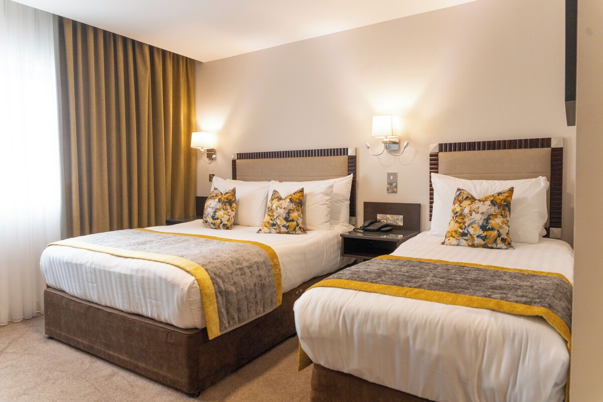 Quarto de hotel com duas camas, cada uma com cabeceira, mesas de cabeceira, abajures e almofadas decorativas.