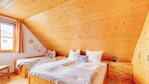 A bedroom with two wooden beds, a wooden ceiling, a window with curtains, and a bedside table with drawers. - Ferienhaus Nora (Sankt Kanzian am Klopeiner See)
