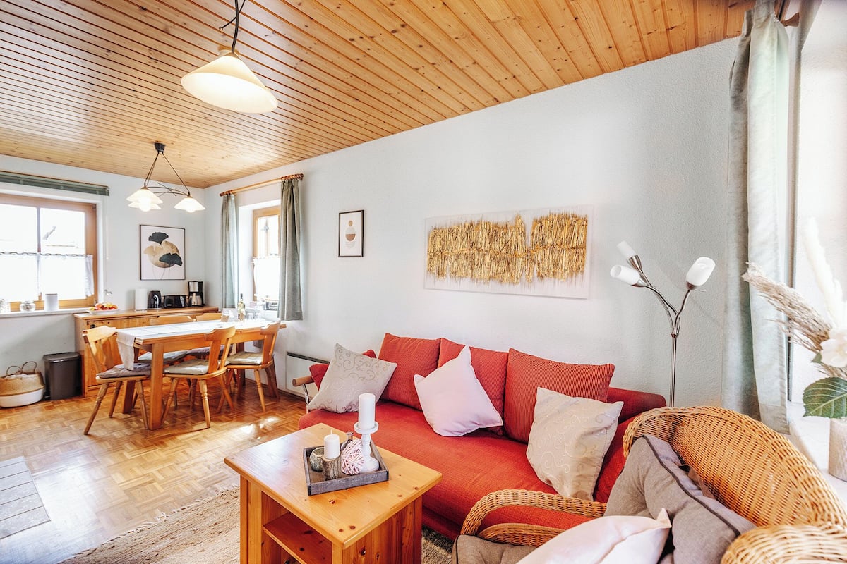 A cozy living room with a wooden ceiling, a dining area with a table and chairs, and a red sofa with cushions.