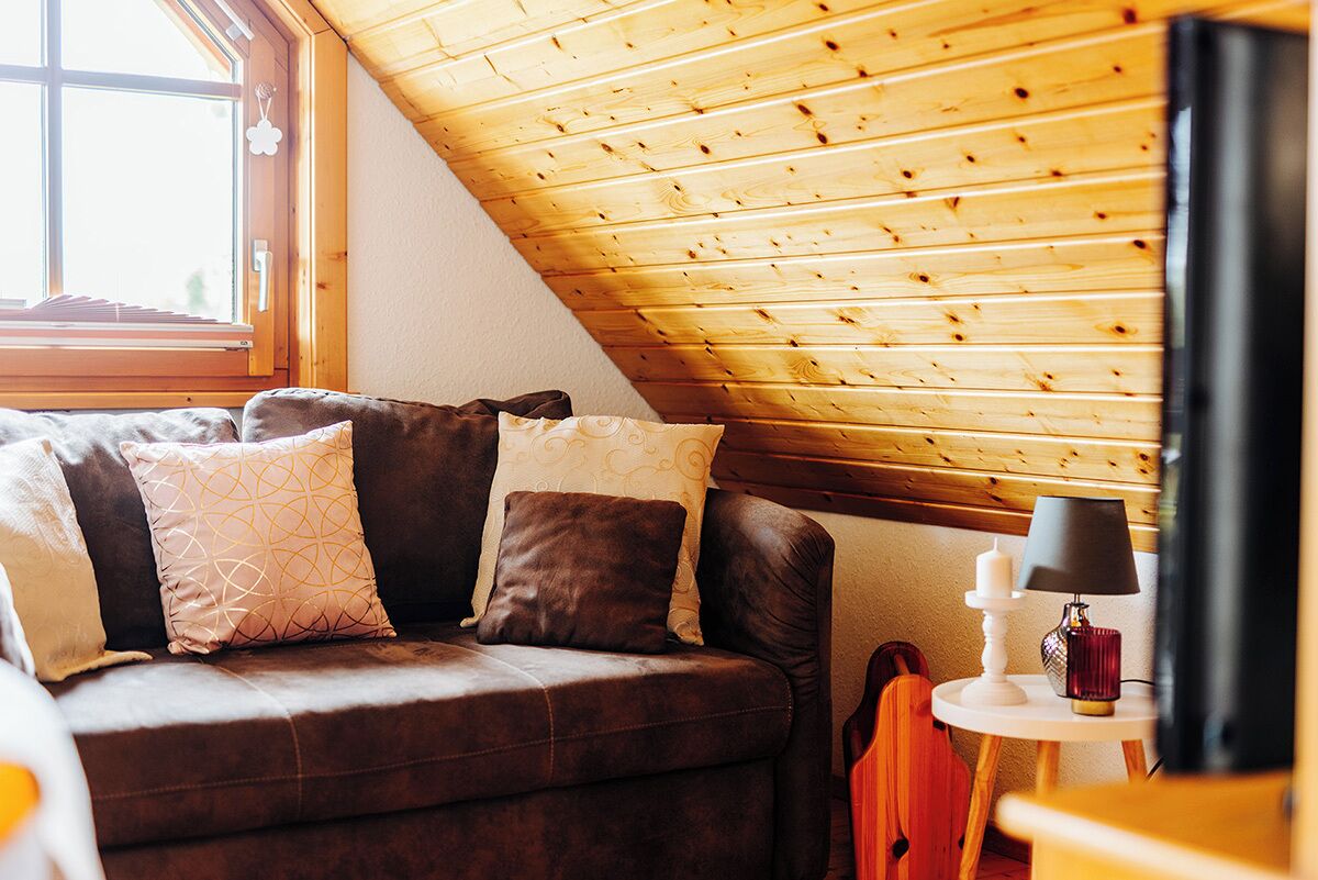 A cozy room with a brown leather sofa, a small round table with a lamp, and a wooden sloped ceiling.