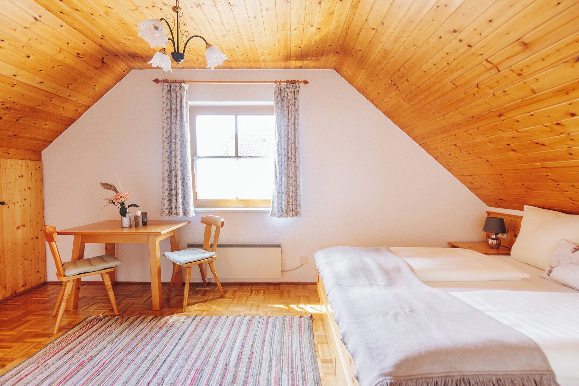 A cozy attic bedroom with a wooden ceiling, a bed, a desk with a chair, and a window with curtains.