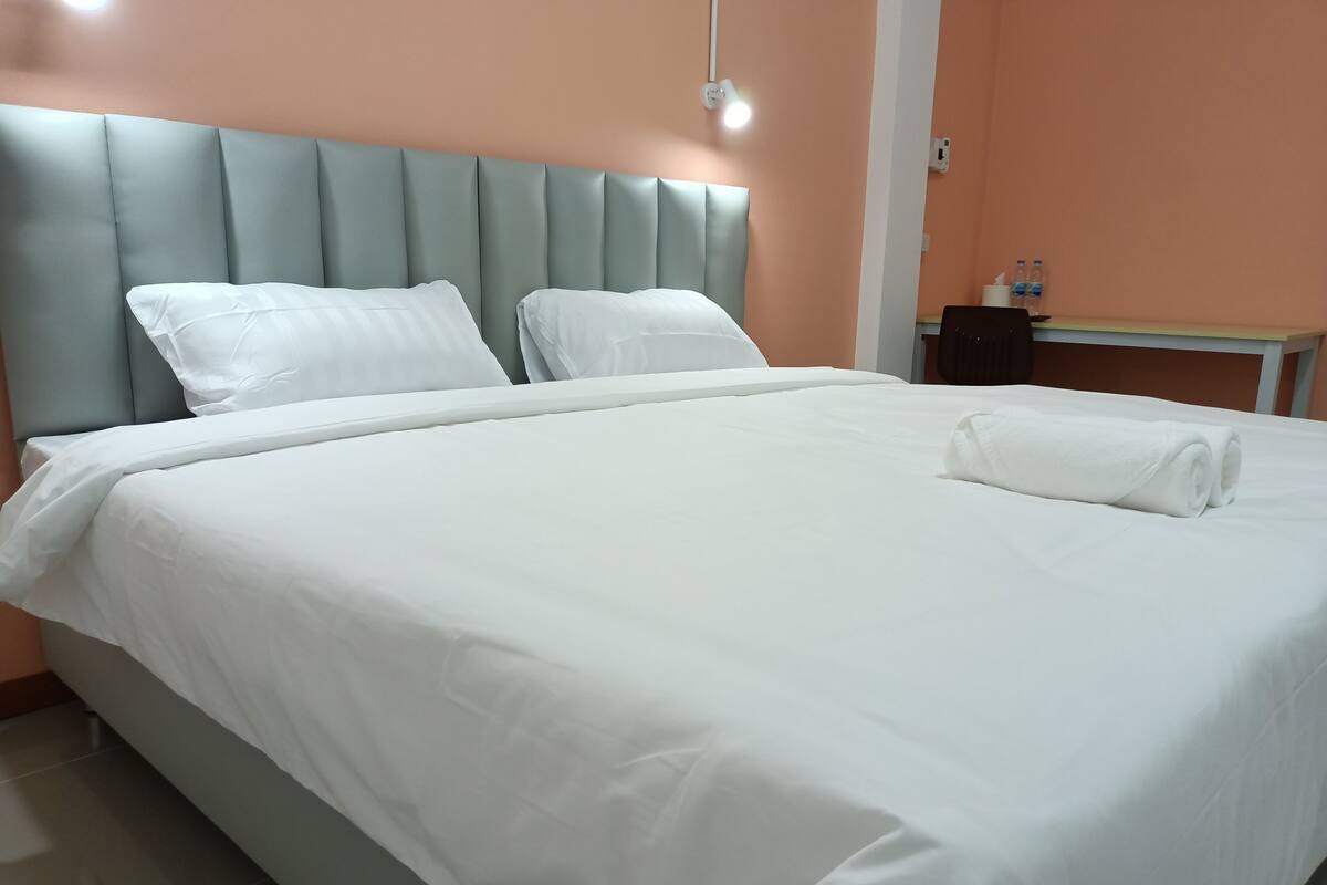 A neatly made bed with white linens, a padded headboard, and a rolled towel on the side.
