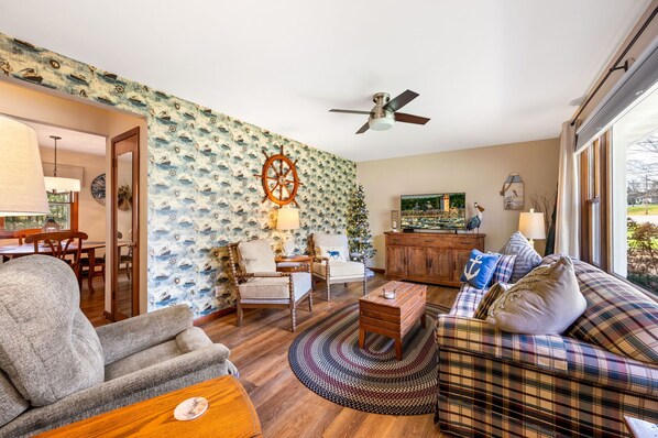 A living room with a nautical-themed wallpaper, a ceiling fan, and a large window with a view of the outdoors. - Drop Anchor | Door County Cozy Ranch Home (Sturgeon Bay)