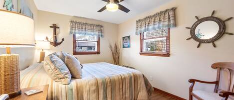 Un dormitorio con cama, una silla, lámpara, ventilador de techo y ventanas con cortinas.