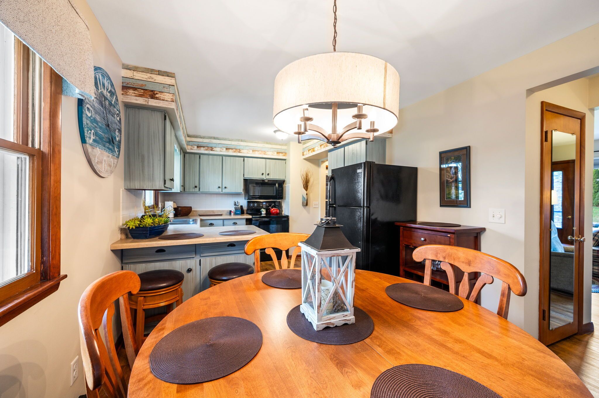 A dining area with a wooden table, chairs, a chandelier, and a kitchen in the background.
