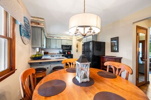 A dining area with a wooden table, chairs, a chandelier, and a kitchen in the background. - Drop Anchor | Door County Cozy Ranch Home (Sturgeon Bay)
