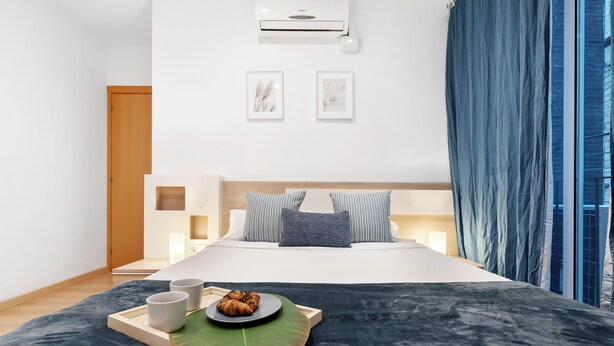 A modern hotel room with a large bed, a bedside table with a lamp, a tray with pastries, and a window with curtains.
