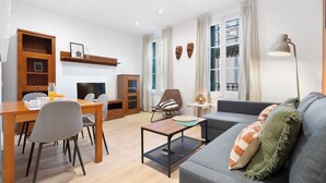A modern living room with a sofa, dining table, and chairs. There is a fireplace, a bookshelf, and a lamp.