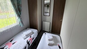 A room with two beds, a radiator, a mirror, and a window with curtains. - Wl34 Caravan Haggerston Castle Private Decking (Berwick-upon-Tweed)