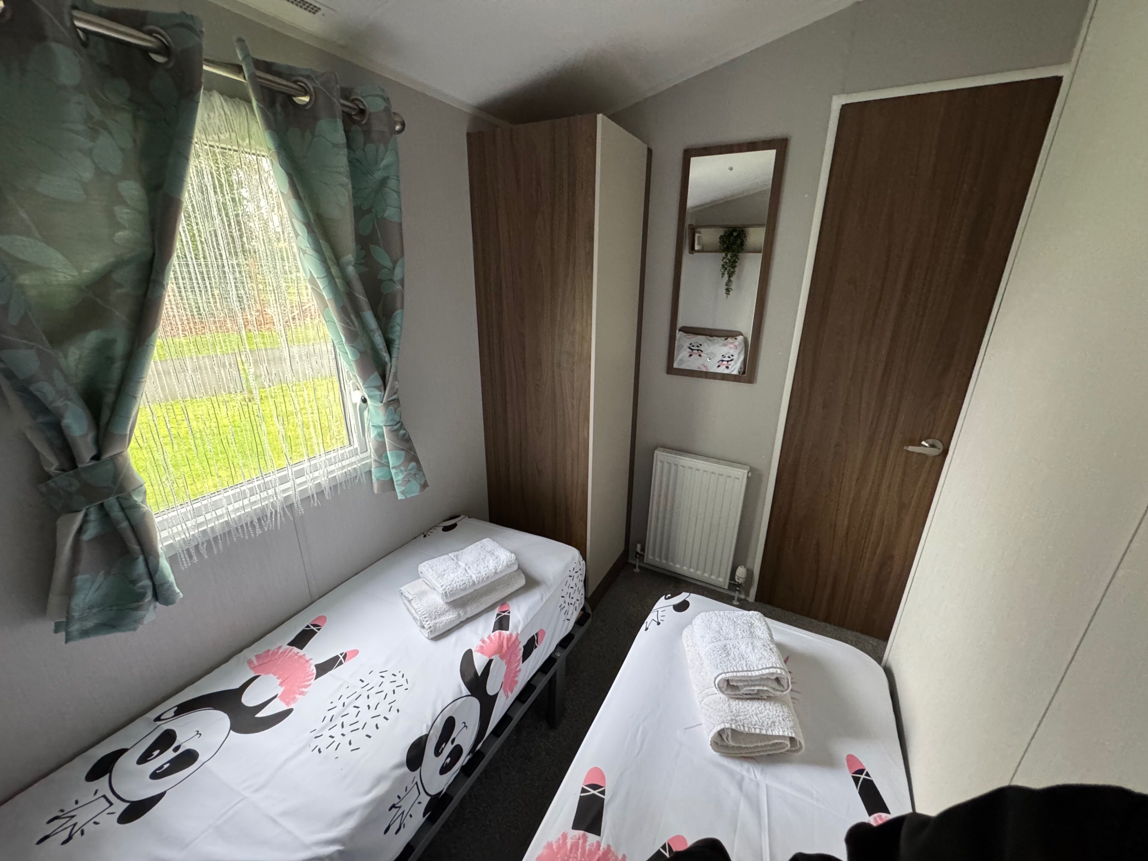 A room with two single beds, a window with curtains, a radiator, and a mirror on the wall.