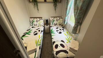 A small room with two beds, each covered with panda-themed bedding. There is a shelf with plants above the beds, and a window with a floral curtain.