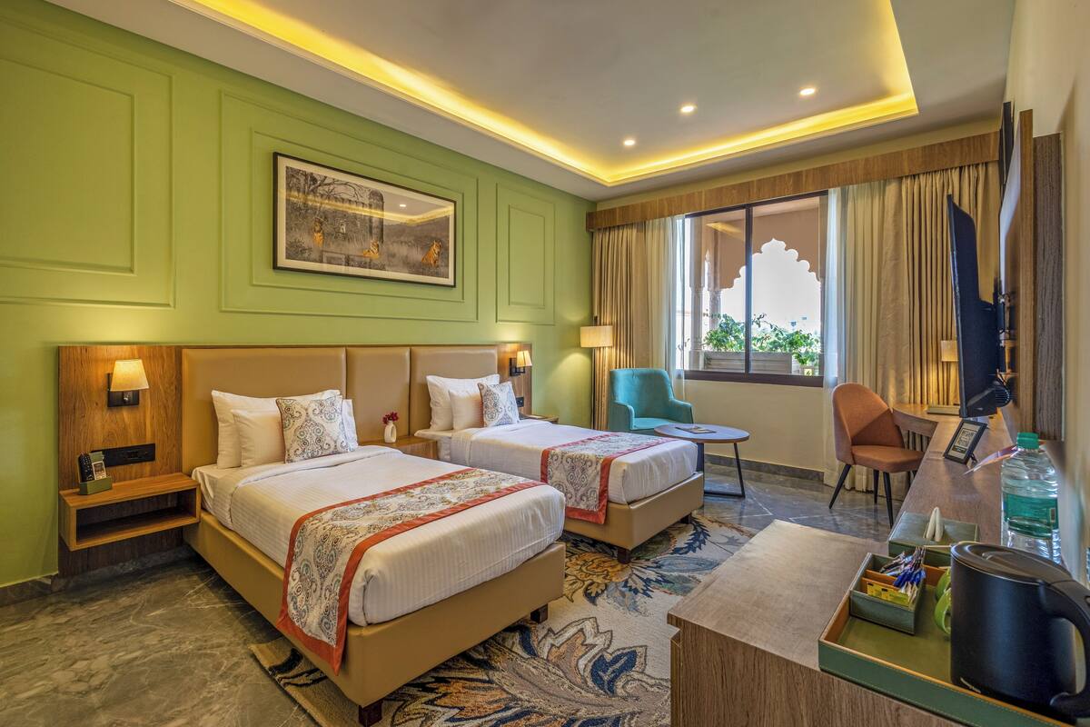 A hotel room with two beds, a desk, a chair, a window with curtains, a painting on the wall, and a small table with a lamp.