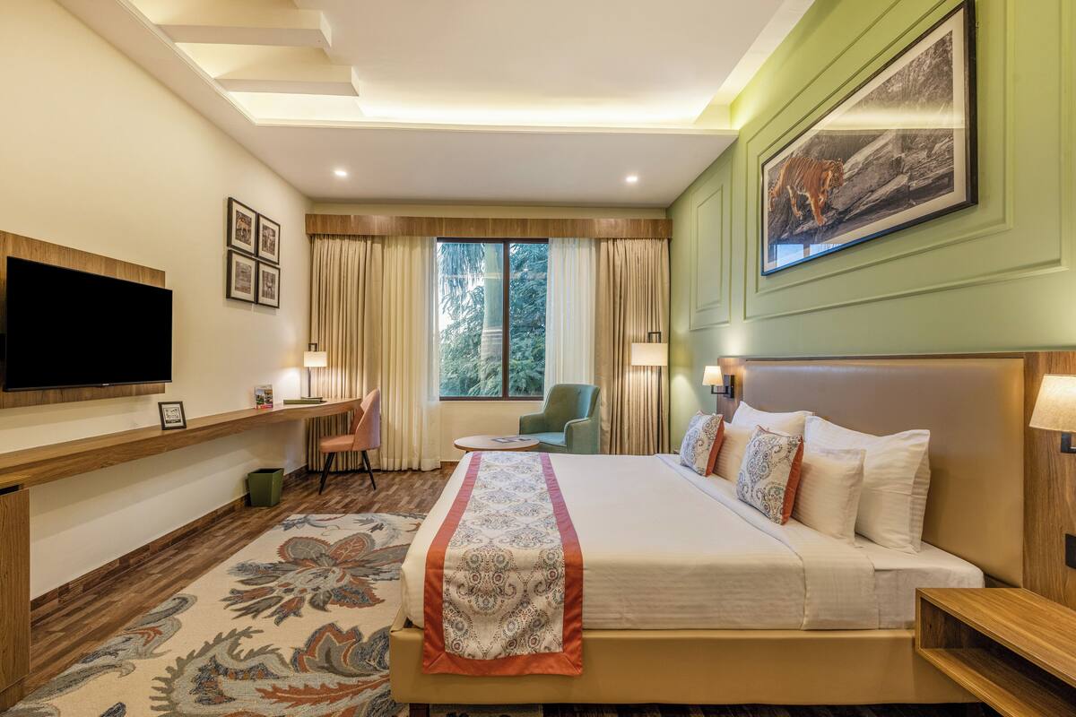 A hotel room with a large bed, a flat-screen TV, a desk with a chair, a small round table, and a window with curtains.