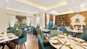 A dining area with tables set for four, green upholstered chairs, and a wooden bookshelf with decorative items. - Sterling Bagh Ranthambore (Sawai Madhopur)