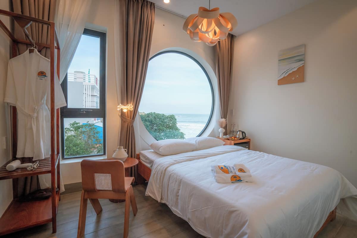 A hotel room with a large round window, a bed with white linens, a wooden chair, a desk with a lamp, and a view of the sea.