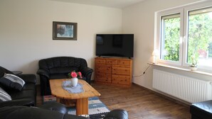 Un salon avec un canapé en cuir noir, une table basse en bois sur laquelle est posé un vase de fleurs, un téléviseur sur un meuble en bois et une fenêtre donnant sur un paysage verdoyant.