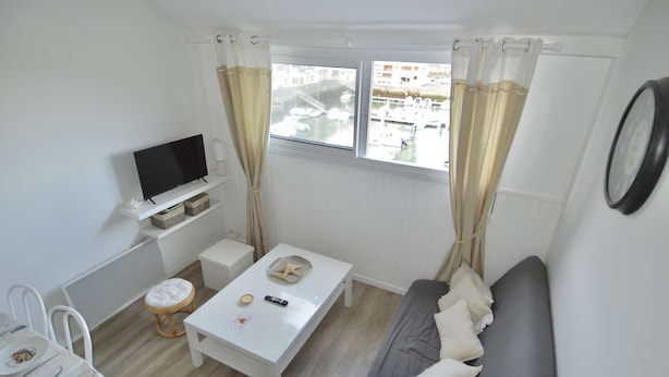 A small, bright room with a TV, a white table, a grey sofa, and a window with curtains. - Aux beaux coquillages, sea and harbor view (Courseulles-sur-Mer)