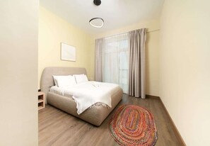 A bedroom with a bed, a bedside table, a window with curtains, and a patterned rug. - Dubai Al Jaddaf Apt Pool Gym Free Parking (Dubai)