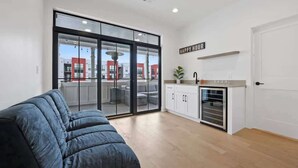 Ein modernes Wohnzimmer mit einer großen Schiebeglaswand, die auf einen Außenbalkon führt, einer Küchenzeile mit weißen Schränken und einem Schild mit der Aufschrift 'Happy Hour'.