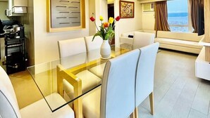 Un espace de salle à manger moderne avec une table en verre, des chaises à revêtement blanc, un éclairage suspendu et un vase de fleurs.
