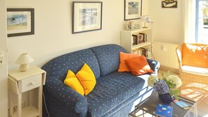 A living room with a blue sofa, two yellow pillows, a white side table with a lamp, a bookshelf, and a wicker chair with an orange cushion. - Quiet, wooded apartment in Eichenpark Prerow, 2 pers., WLAN, south-west-facing balcony (Ostseebad Prerow)