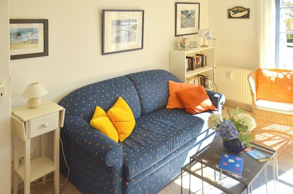 Een woonkamer met een blauwe bank, twee gele kussens, een witte bijzettafel met een lamp, een boekenkast en een rieten fauteuil met een oranje kussen.