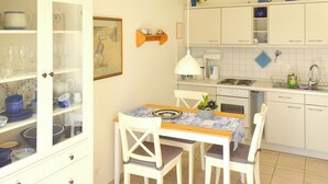 A small kitchen with white cabinets, a dining table with chairs, and a hanging light. - Quiet, wooded apartment in Eichenpark Prerow, 2 pers., WLAN, south-west-facing balcony (Ostseebad Prerow)