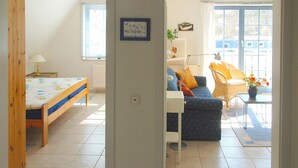 A room with a bed, a sofa, a small table with flowers, and a window with blinds. - Quiet, wooded apartment in Eichenpark Prerow, 2 pers., WLAN, south-west-facing balcony (Ostseebad Prerow)