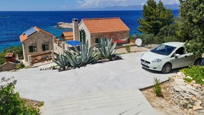 Egy tengerparti ingatlan, melyhez aszfaltozott felhajtó vezet, egy fehér autó áll előtte, és egy kőből épült épület látható, vörös tetővel.