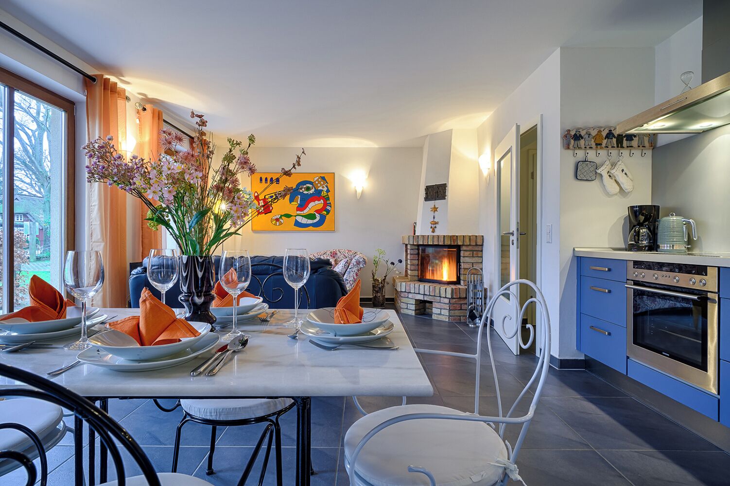 Un coin repas avec une table dressée pour quatre personnes, une cheminée et une cuisine équipée d’appareils électroménagers modernes.