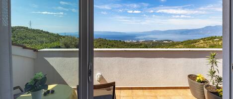 Un balcón con mesa y sillas, una planta en maceta y vistas a un paisaje costero con colinas y el mar.
