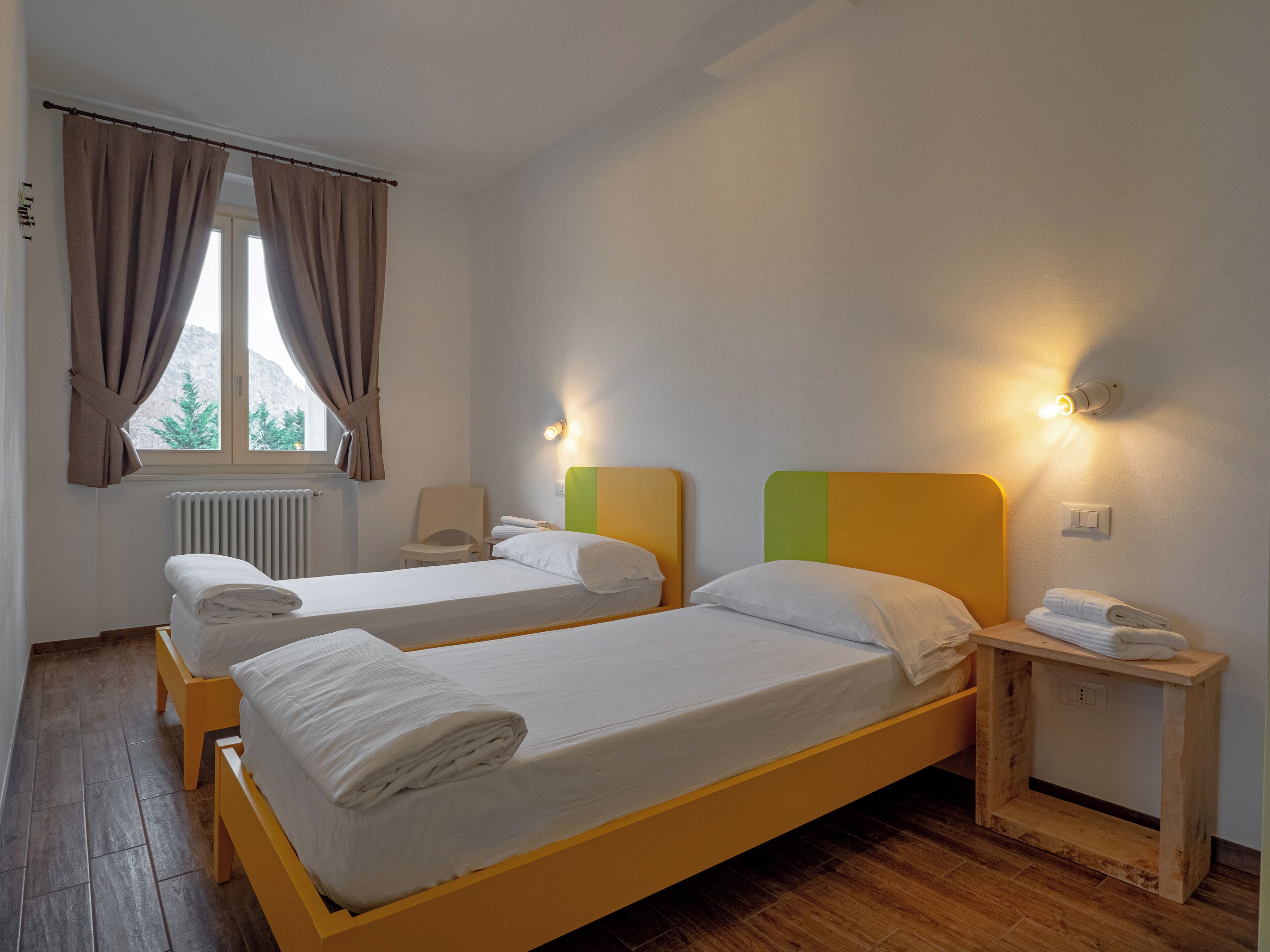 Ein Hotelzimmer mit zwei Einzelbetten, einem Holfnachtkästchen, einem Stuhl und einem fenster mit Vorhängen.